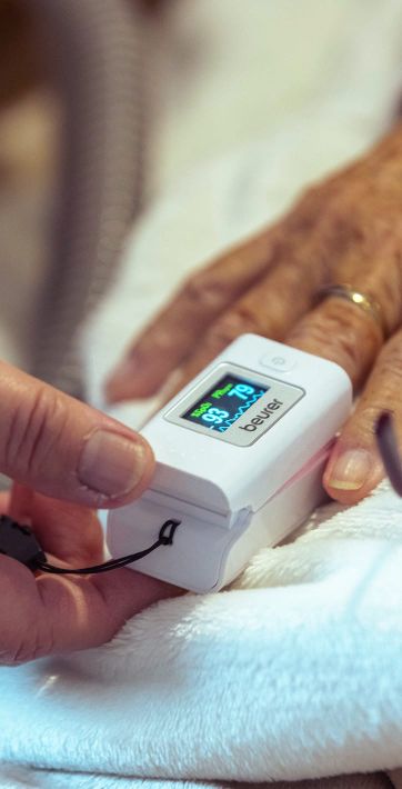 Eine Hand legt ein Puls-Oximeter auf einen Finger, während die Werte auf dem Display angezeigt werden.