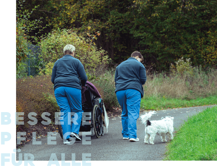 Zwei Pflegekräfte begleiten einen Rollstuhl auf einem Weg, während ein Hund neben ihnen herläuft.