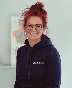 Korinna Grebe lächelt freundlich in einem Akademed-Hoodie vor einer Wand mit anatomischem Poster.