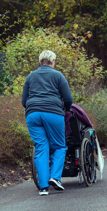 Eine Pflegekraft schiebt einen Rollstuhl entlang eines schmalen Weges, umgeben von Herbstlaub.