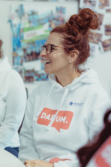 Eine Frau in einem weißen Hoodie mit der Aufschrift "DARUM." lächelt während einer Besprechung.