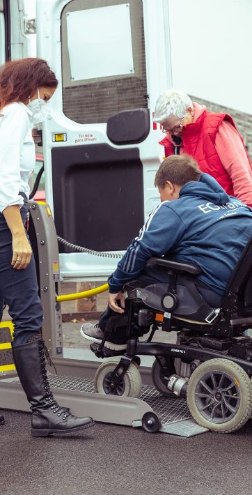 Eine Frau hilft einem Jungen im Rollstuhl, während eine ältere Frau beim Einsteigen in ein spezialisiertes Fahrzeug assistiert.