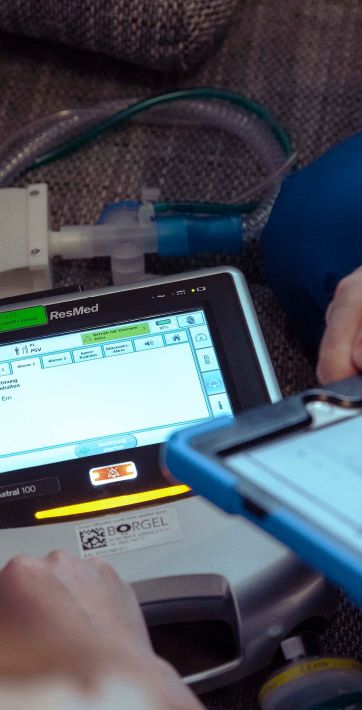 Eine Person bedient ein medizinisches Gerät von ResMed zur Unterstützung der Atemtherapie, während ein Tablet in der Hand gehalten wird.