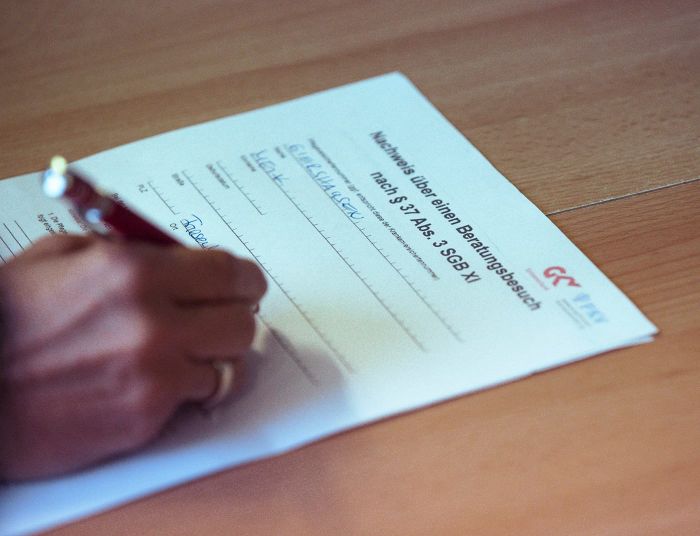 Eine Hand füllt ein Formular für einen Beratungsbesuch nach § 37 Abs. 3 SGB XI auf einem Tisch aus.
