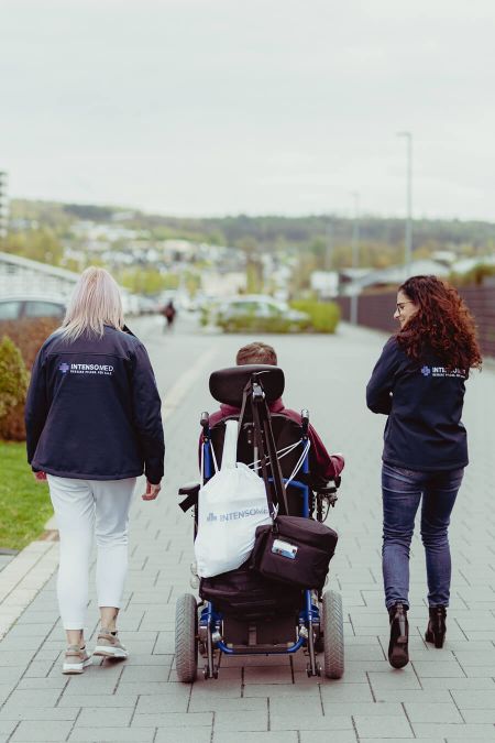 Drei Personen, darunter eine Person im Rollstuhl, spazieren auf einem gepflasterten Weg in einer grünen Umgebung.