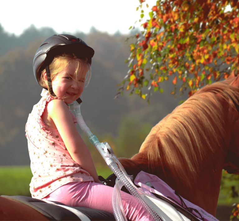 Ein kleines Mädchen mit einem Helm lächelt glücklich auf einem Pferd, während es eine Atemhilfe verwendet.