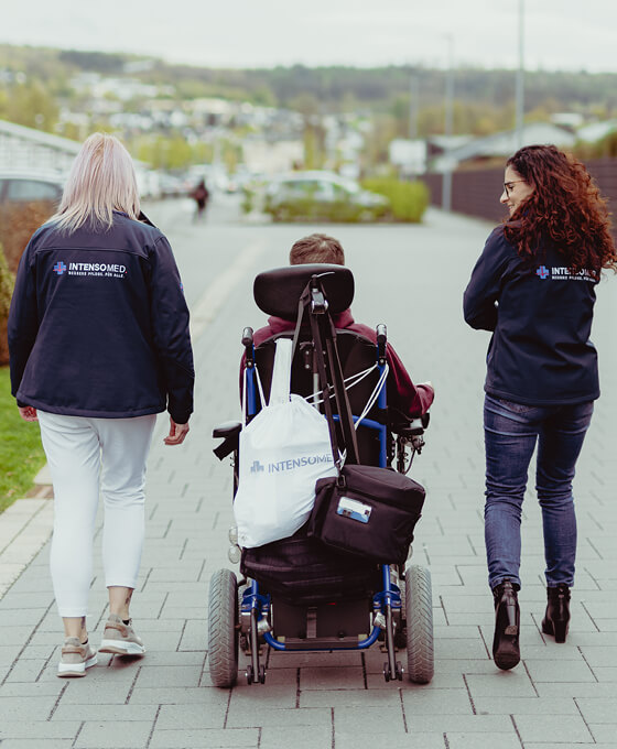 Drei Personen, darunter eine Person im Rollstuhl, spazieren auf einem gepflasterten Weg in einer grünen Umgebung.